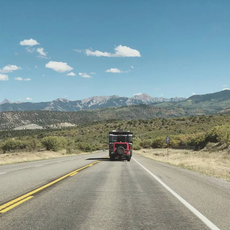 An open road towards mountains, behind an RV towing a red Jeep